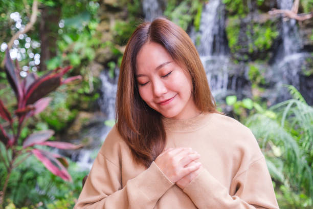 porträtbild einer jungen frau mit geschlossenen augen, die im garten die hände auf ihre brust legt - spirituality hand on heart meditating women stock-fotos und bilder