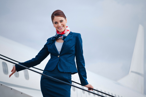 Outdoor portrait of a beautiful flight attendant standing on the aircraft stairs and smiling at the camera.