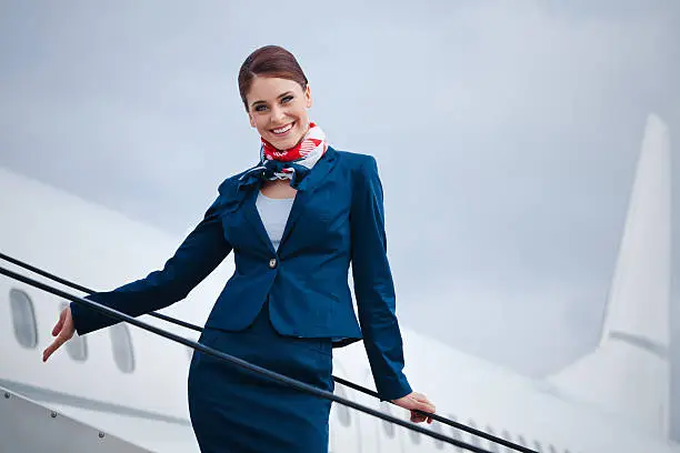 Outdoor portrait of a beautiful flight attendant standing on the aircraft stairs and smiling at the camera.