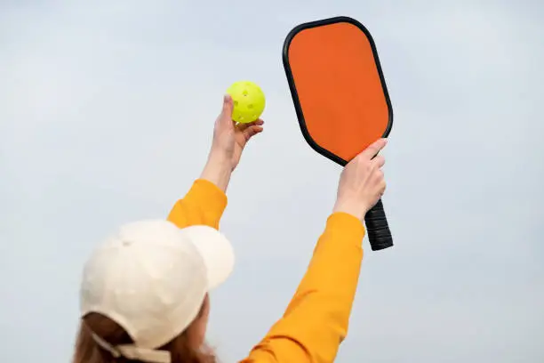 Photo of back view pickleball game woman player, hands over blue sky hitting pickleball yellow ball with paddle, outdoor sport leisure activity.