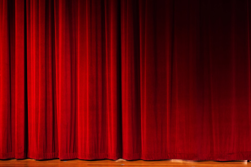 Closed red velvet stage drapes, lit by footlights, await the performer, speaker, or event. The floor of the stage is just visible. Also useful as graduated red, textured background.