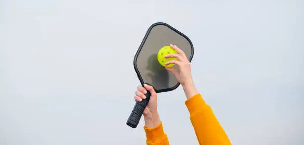 Photo of banner pickleball game, hands over blue sky hitting pickleball yellow ball with paddle, outdoor sport leisure activity.