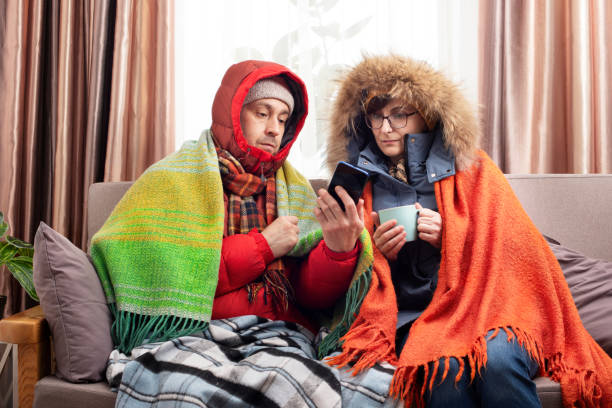 istock Middle age family in parkas drinking coffee on the sofa while using smartphone in their cold living room 1685891164