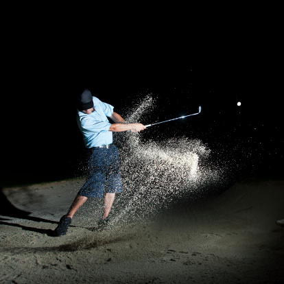 A golfer hitting out of a sand bunker at night with stars above.