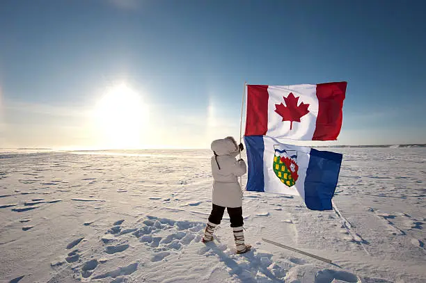 A sundog rises from the horizon at noon on a cold January day on Great Slave Lake near the city of Yellowknife in canada's Northwest Territories.  A woman in winter parka holds the Canada and Northwest territories flags in a brisk wind.  Copy space image left.