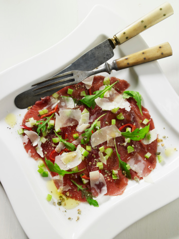 White plate with serving of bresaola and parmesan cheese, sprinkled with salt, pepper, olive oil,rucola (Eruca sativa) and spring onion. Antique set of knife and fork on the side