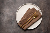 Minimal table setting with beige plate, brown linen napkin and gold cutlery, brown rustic background. Top view, flat lay.