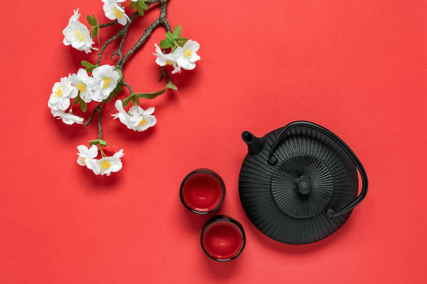 アジアの春の茶道。赤い背景に黒い鋳鉄製のティーポット、カップ、装飾的な花の咲く桜の枝。上からの眺め。