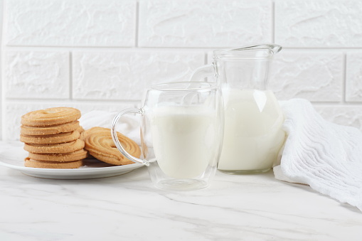 Milk in a transparent glass and jar on a marble table. Round ring shaped German spritz biscuits at background