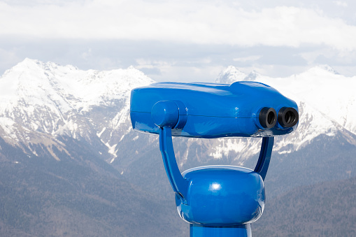 Blue public binoculars in mountains. Tourist telescope against blurred mountain peaks background. Binoculars for sightseeing.