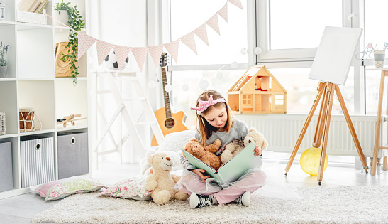 Happy little girl reading book with plush teddies in light room