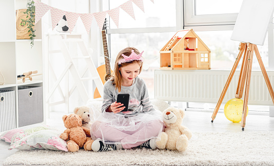 Lovely little girl holding phone sitting on floor with plush teddies