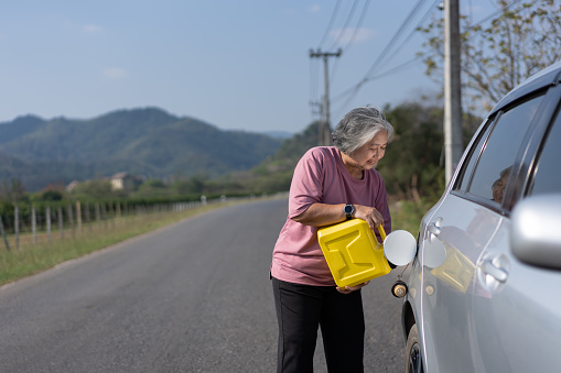 The car ran out of gas and stalled beside the road in suburbs and an elderly Asian woman used a gallon of spare gas to fuel the car. A woman prepares a gallon of spare gas to fuel before traveling.