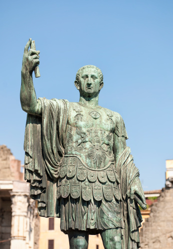 A heavily weathered bronze statue of a Roman Emperor holding a scroll, situated near Trajan's Forum on Via dei Fori Imperiali in central Rome.