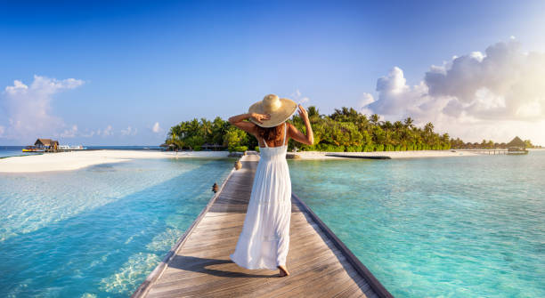 Back view of a woman in white dress walking down a pier towards a tropical island Back view of a elegant woman in white dress and hat walks down a pier towards a tropical island in the Maldives floating dock stock pictures, royalty-free photos & images