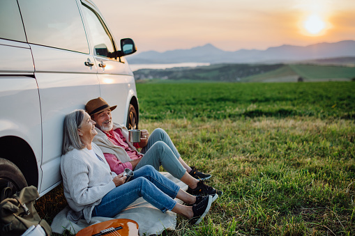 Potrait of senior couple sitting by ste car and drinking coffee after long drive during their roadtrip. Elderly spouses at roadtrip enjoying serene landscape of High Tatras behind them.