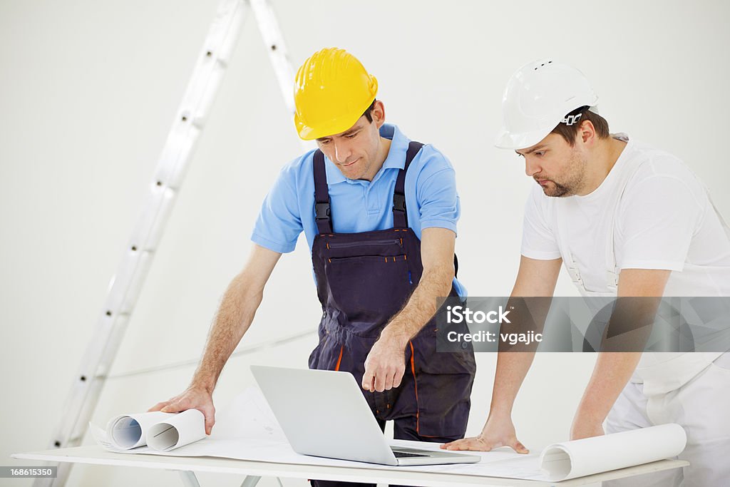 Manual Workers At Construction Site Manual workers with protective workwear expertise project on laptop and blueprint. House Painter Stock Photo