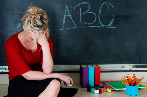 Elementary classroom setting with tired or frustrated teacher holding her head. She's sitting in front of an chalkboard with ABC
