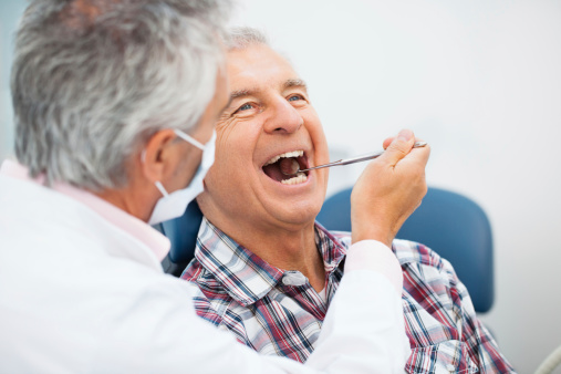 A gray-haired patient opens his mouth while slightly reclined in a blue chair at a dentist's office.  A gray-haired dentist seated next to him holds an examination tool in the patient's mouth. The patient is wearing a plaid shirt, and the dentist is wearing a white coat and mask.