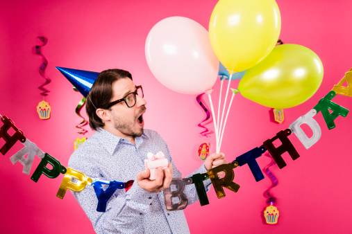 Happy birthday! Surprised Geek man with horn rimmed glasses, blue party hat, gift and five party multicolored decoration balloons in hand, isolated on pink background.