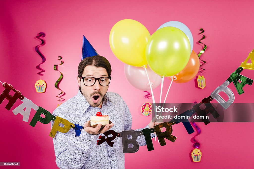 Surprised Nerdy man holding gift and multicolored balloons, Happy birthday! Happy birthday! Surprised Geek man with horn rimmed glasses, blue party hat, gift and five party multicolored decoration balloons in hand, isolated on pink background. Arts Culture and Entertainment Stock Photo