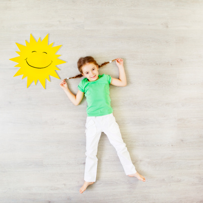 Cute little girl lying on laminate floor with foam sun, presenting sunny weather.