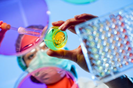 Image of a chemist working at a hospital lab while wearing protective gear. Around the researcher we have a microtiter plate and a petri dishes while she is holding a volumetric flask and a dropper. Image created with very shallow depth of field. Focus point is on her fingers and volumetric flask.