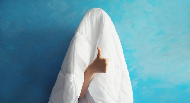 Cheerful lazy woman waking up after sleeping covered head with white blanket showing cool gesture her fingers on blue background stock photo