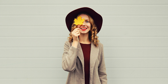 Autumn color style outfit, portrait of stylish happy smiling young woman covering her eyes with yellow maple leaves wearing hat on gray background