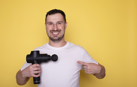 Young man wearing white clothes spend time in home gym hold and pointing on massage gun isolated on yellow background.