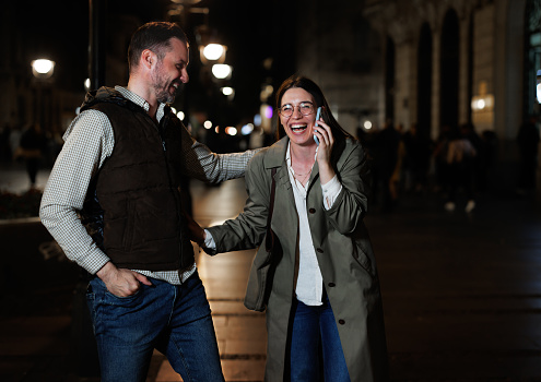 Under the shimmering city lights, a couple deepens their bond as they embark on a digital journey together, engrossed in their mobile phones, exploring, sharing, and discovering the digital world while creating cherished memories along the bustling city street