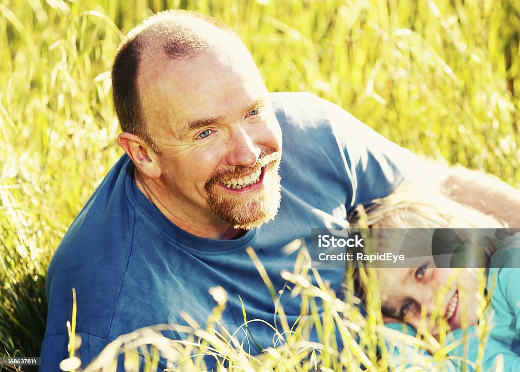 Father and little blonde daughter relaxing together in field A pretty little blonde girl smiles  in delight as she cuddles up with her father in long grass in a field. 4-5 Years Stock Photo