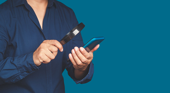 Search online concepts. A businessman is holding a magnifying glass and looking at a smartphone while standing on a blue background. Space for text