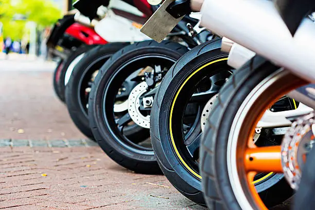 A row of motorcycles with differing wheels and tyres lined up on the diagonal on a brick-paved surface