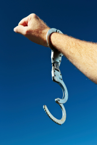A man's hand, holding up a clenched fist in a salute, and wearing a single, open handcuff, is brandished against clear blue sky. Symbolic of release or freedom from almost anything! 