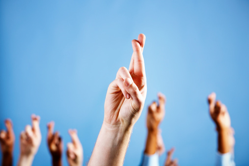 Many hands are raised against a sky blue background, all with fingers crossed superstitiously, just in case! Focus is on the man's hand in the foreground. 