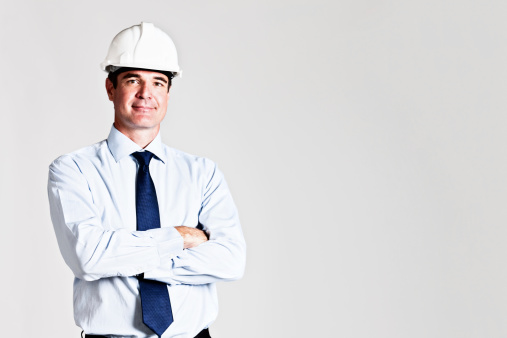A handsome, confident-looking man in a hard hat and a formal business suit with folded arms looks at the camera, smiling. He could be a construction boss, a civil engineer or an architect.