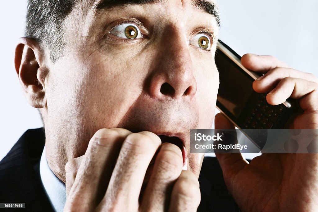 Shocked businessman biting nails in panic at phone call A good looking mature businessman is shocked, biting his nails in panic at something he hears on his mobile phone, bad news or possibly a threatening phone call! Terrified Stock Photo