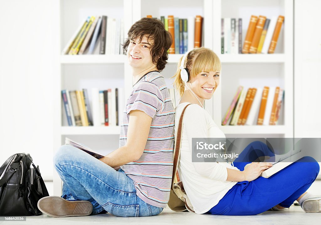 Students in a library. Portrait of an young couple reading book in a library. Sitting on floor in a library and looking at camera. 16-17 Years Stock Photo