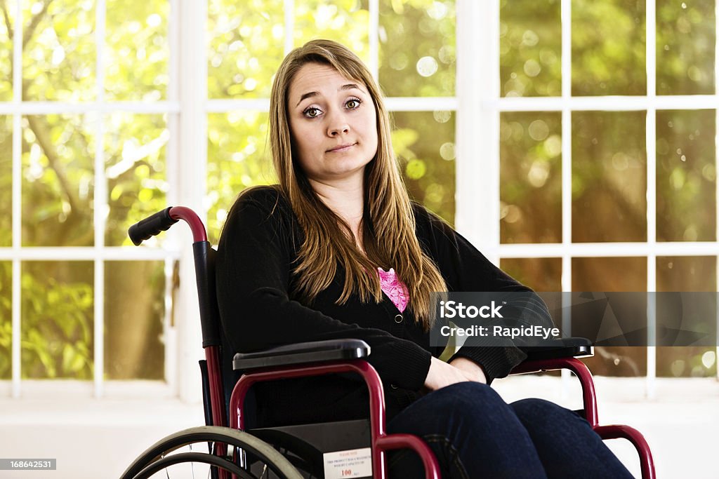 Resigned young woman in wheelchair shrugs as if saying &quot;Whatever&quot; A beautiful young woman sits in her wheelchair looking resigned but mildly irritated, her eyebrows raised and shrugging,  frustrated by her situation and possibly in pain. 20-29 Years Stock Photo