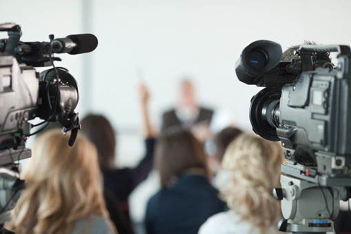 Video cameras pointed at businessman or lecturer giving a presentation in background. Focus on cameras.  