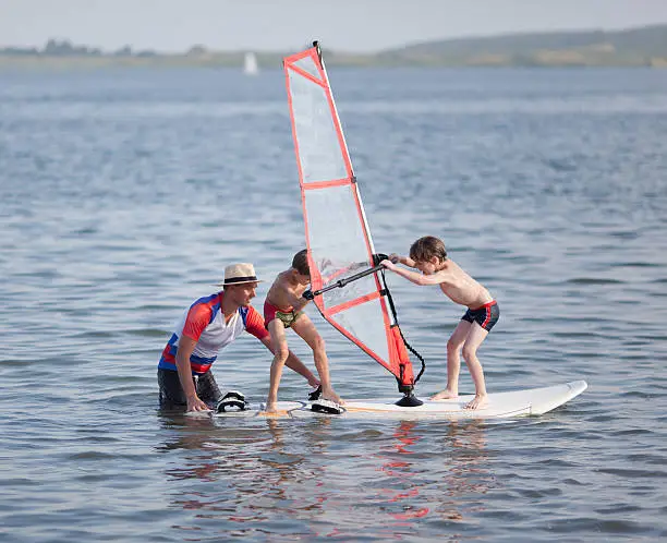 Two little boys try to balance on windsurfing board  with child's sail.  Next to them is male instructor