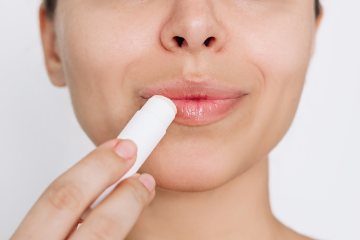 Cropped shot of a young caucasian attractive woman applying a hygienic lipstick on her lips on a light gray background. Moisturizing chapstick for dry lips