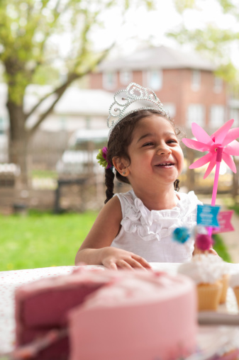 Young mixed-race girl laughing wearing a birthday tiara and holding a pinwheel in her backyard. Out of focus birthday cake and cupcakes in the foreground.