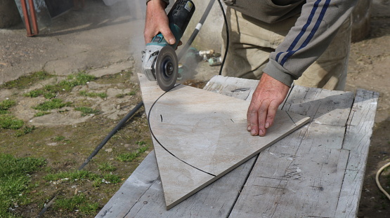 cutting large ceramic tiles with a grinder along a black oval outline in an outdoor tiler's workshop, trimming finishing tiles with a miter saw to the required size