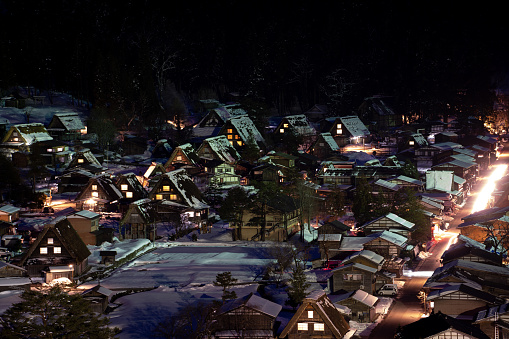 Under the veil of night, the winter-enshrouded Shirakawa village illuminates with the soft glow of lighted houses. The snow blankets the rooftops, reflecting the warm amber tones of the interior lights. Beyond the homes, the darkness is punctuated by the silent shadows of trees and distant mountains. This nighttime tableau evokes a serene and magical ambiance, capturing the timeless charm of a winter evening in rural Japan.