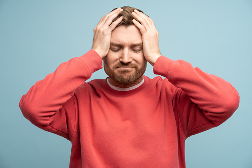 Irritated man with closed eyes holds head feeling discomfort due to headache on blue wall background. Concept of illness and suffering from unpleasant sensations. Sad bearded male regrets a mistake