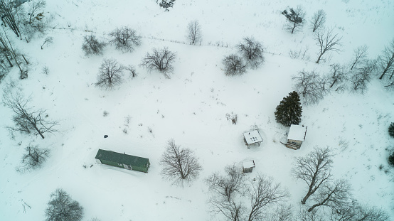 An aerial drone shot of a green sea container and a tiny off-grid log cabin in a field of snow by the woods during winter