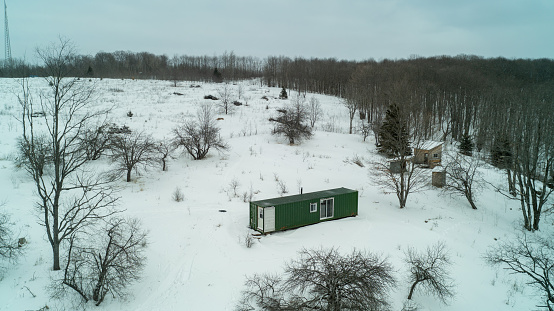 An aerial drone shot of a green sea container and a tiny off-grid log cabin in a field of snow by the woods during winter