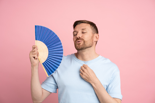 Portrait of playful tired man waving blue fan suffers from stuffiness standing on pink background. Overheating high temperature, hot summer weather concept. Banner, poster of posing artistic male.